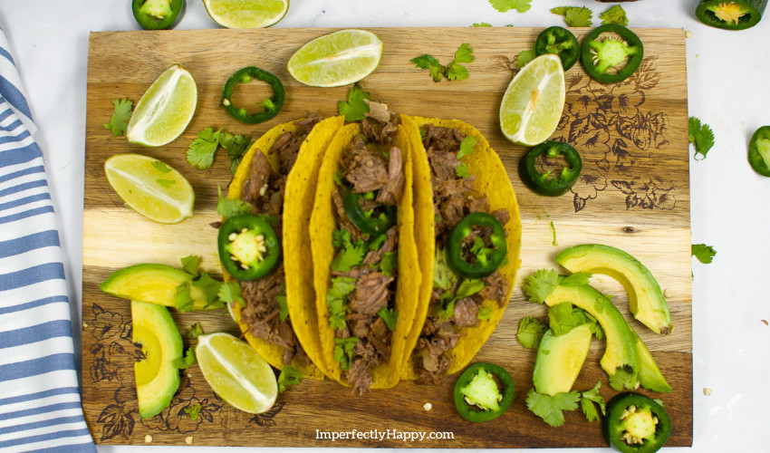 Beef Barbacoa meat in crispy taco shells topped with fresh jalapenos on a wooden cutting board. Tacos are surrounded by limes and avocado.