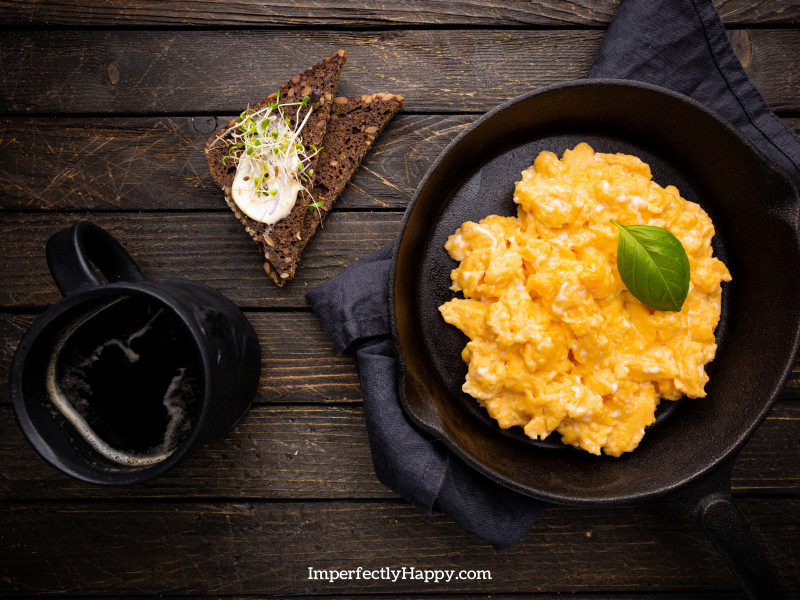 Scrambled eggs in cast iron skillet on wooden table with mug of black coffee and toast