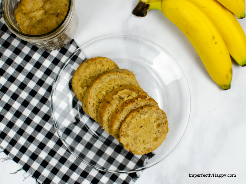 Banana bread recipe - bread sliced on clear plate. Banana bread in a jar and bananas in the background. Black and white checkered cloth under plate.